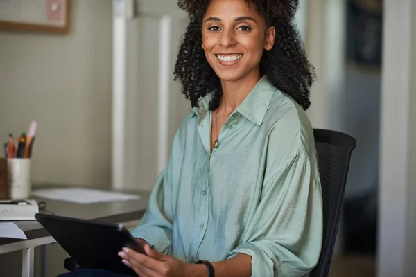 Portrait of a smiling young African female entrepreneur working on a digital tablet while sitting in a chair in her home office