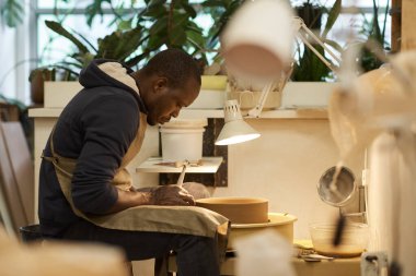 Young male African pottery maker shaping a bowl spinning on a wheel using a handle maker tool in a ceramics studio clipart