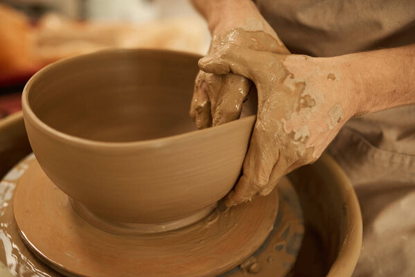 Close-up of a male potter shaping a piece of clay into a bowl with his wet hands on a pottery wheel in a ceramics studio