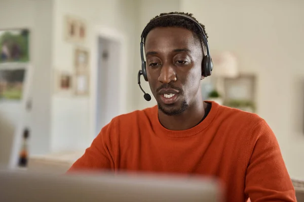 Young African male entrepreneur wearing a headset and talking on a video call on a laptop while working remotely from home