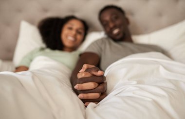 Closeup of an affectionate young multiracial couple smiling and holding hands together while lying in bed in the morning