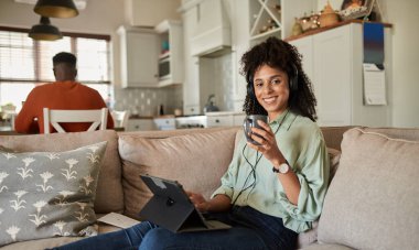 Gülümseyen Afrikalı kadın kulaklık takıp kahve içiyor ve kocası mutfakta otururken kanepede tablet kullanıyor.