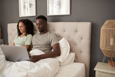 Genç, çok kültürlü bir çift sabahları yataklarında oturup dizüstü bilgisayar ve dijital tablet kullanıyorlar.