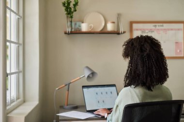 Genç bir Afrikalı kadın girişimcinin dikiz aynasından bilgisayarla çalışırken ev ofisindeki bir masada oturması.