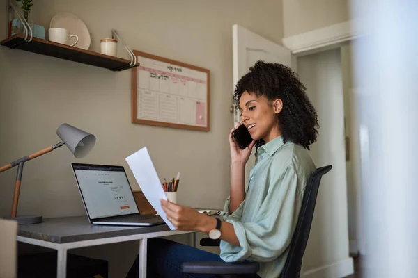Young African female entrepreneur talking on a phone while reading paperwork and working on a laptop at a desk in her home office