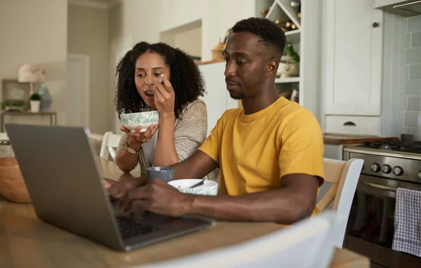 一对年轻的多种族夫妇在厨房的一张桌子旁 一边吃早餐 一边上网浏览笔记本电脑 — 图库照片
