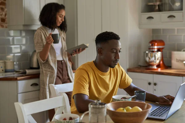 年轻的多种族夫妇在家里的厨房边喝咖啡边吃早餐边看笔记本电脑 — 图库照片