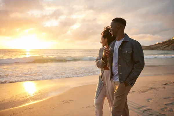 砂浜で腕を組んで歩いている間に夕日を見ている若い多民族のカップルを笑っています — ストック写真