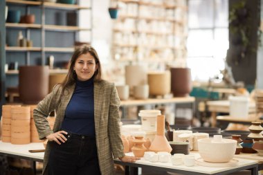 Gülümseyen genç bir kadın seramik stüdyosu sahibinin portresi. Parlak bir çömlek imalat stüdyosunda çalışma tezgahlarının yanında duruyor.