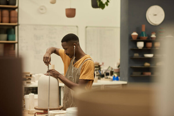 Молодой африканский керамист, работая на скамейке в керамической мастерской, делает дыру в большом глиняном горшке