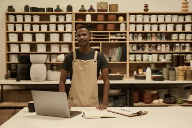 Büyük bir seramik stüdyosunun çalışma masasında dizüstü bilgisayarın başında duran gülümseyen genç bir Afrikalı çömlekçinin portresi.