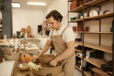 Seramik bir stüdyoda, yeni yontulmuş bir kaseyi çömlek çarkına koymak için kablo kullanan genç bir erkek seramik.