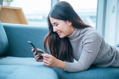 Kanepede oturan kadınlar akıllı telefon, internetten alışveriş yapmak için kredi kartı ve alışveriş düşkünleri için mutlu yüzler kullanıyorlar. Mutlu Asyalı kadınlar akıllı telefonlarıyla uzanıyorlar. Kadın online alışveriş ödemesi