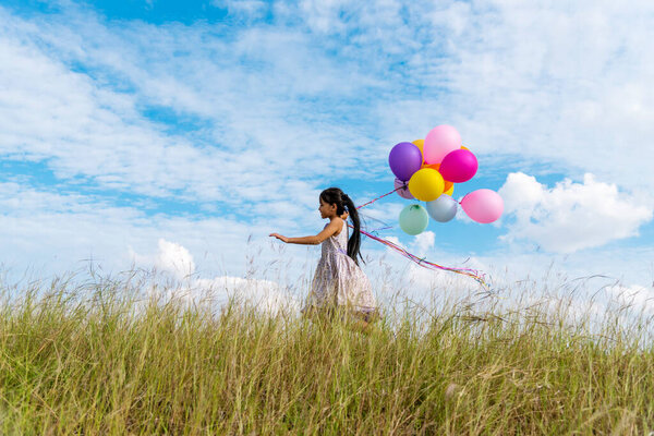 Cheerful cute girl holding balloons running on green meadow white cloud and blue sky with happiness. Hands holding vibrant air balloons play on birthday party happy times summer on sunlight outdoor