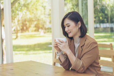 Asyalı kadın, yeşil bahçe kafesinde elinde fincanla sade kahve içiyor. Genç kadın gülümsüyor, aşk gülüyor, kahve içiyor. Kahve dükkanında elinde taze fincanla sade kahve içen güzel kadın. Kahve sever.