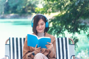 Kitap okuyan mutlu Asyalı kadın kulaklık takıyor, açık havada müzik dinliyor yeşil park doğasında. Kitap okuyor, rahatlama için müzik terapisti. Yeşil parkta oturan mutlu kadın gülüşü