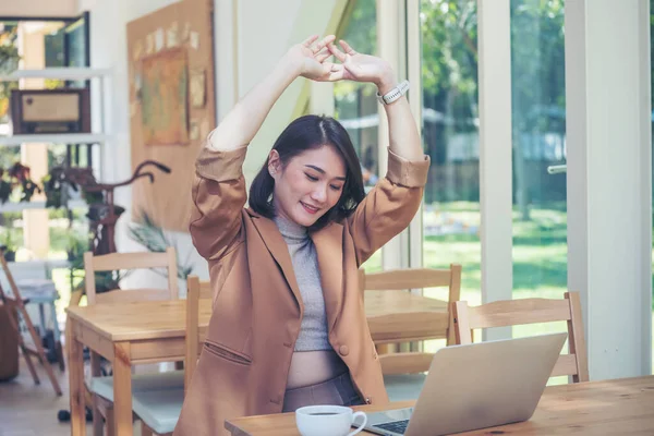 Woman stretching arm raised sitting incorrect position at home office desk. Young asian woman tired from work body stress back pain office syndrome. freelance Female work from anywhere using laptop