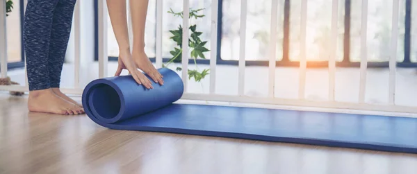 Banner Woman hands rolled up yoga mat on gym floor in yoga fitness training room. Home workout woman hands rolling foam yoga gym mat background. Panoramic Woman barefoot home workout with copyspace