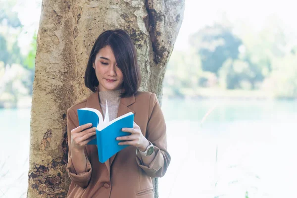 Happy asian woman reading book outdoor in green park nature. Reading book magazine relaxation woman. Leisure portrait woman smile sitting in green park read e-book diary planner with happy face