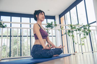 Asana yogası spor kıyafetleri içinde gülümseyen güzel bir kadın yoga minderinde oturup sağlıklı yaşam tarzını seyrediyor. Gülümseyen Asyalı kadın, Asya yoga vücudu, oturma odasında bacak esneme sporu yapıyor.