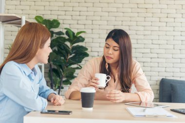 İki bayan meslektaşım ofis masasında kahve fincanı içiyorlar. Asyalı bir kadın arkadaşça sinsice gülüyor. Takım çalışması mutlu an yaşam tarzı. Uluslararası Kadın Günü konsepti