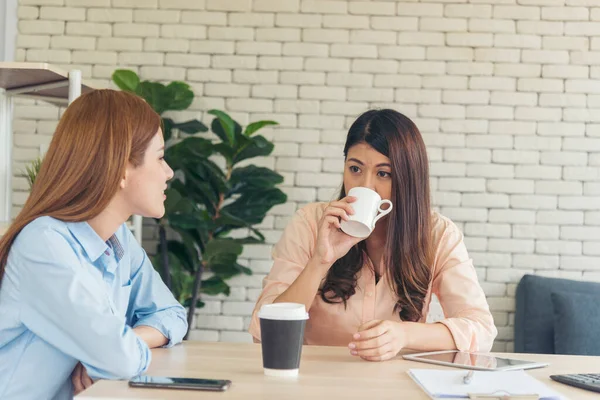 Two Women colleagues drinking black coffee cup at office desk together. Asian woman friendstalking smiling with friendship happy time. Teamwork happy moment lifestyle. International Woman day concept