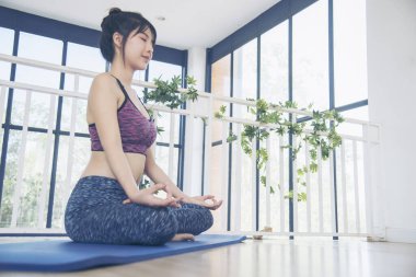 Asana yogası spor kıyafetleri içinde gülümseyen güzel bir kadın yoga minderinde oturup sağlıklı yaşam tarzını seyrediyor. Gülümseyen Asyalı kadın, Asya yoga vücudu, oturma odasında bacak esneme sporu yapıyor.