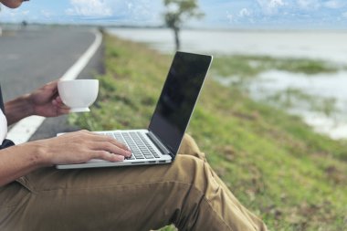 Ofisin dışında elinde kahve fincanıyla dizüstü bilgisayar yazan bir adam. Bilgisayar kullanarak ellerini kapat. Yeşil Park 'ın açık hava yaşam tarzını izle. Laptop ev ofis teknolojisi kullanan serbest çalışan adamlar.