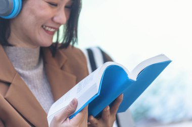 Kitap okuyan mutlu Asyalı kadın kulaklık takıyor, açık havada müzik dinliyor yeşil park doğasında. Kitap okuyor, rahatlama için müzik terapisti. Yeşil parkta oturan mutlu kadın gülüşü