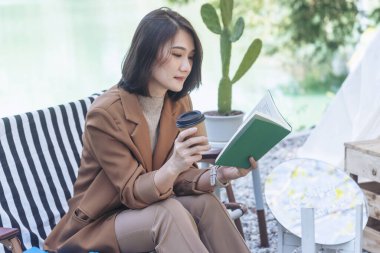 Yeşil Park Doğa 'da kitap okuyan mutlu Asyalı kadın. Kitap okuyarak rahatlayan kadın. Yeşil Park 'ta oturmuş gülümseyen, neşeli yüzlü bir kadın portresi okuyan, e-kitap planlayıcısı.
