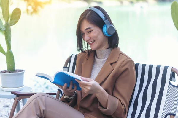 Happy asian woman reading book use headphones listen to music outdoor in green park nature. Reading book magazine, Music therapist for relaxation woman. Happy woman smile sitting in green park
