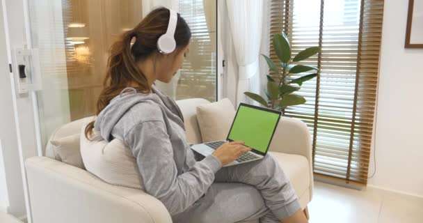ハッピーアジアの女性は 家庭の居間で居心地の良いソファーに座って音楽オンラインWebカンファレンスを聞くヘッドフォンを使用しています オンラインEラーニングビデオ通話を見ながらホームリサーチで働く女性アジア人 — ストック動画