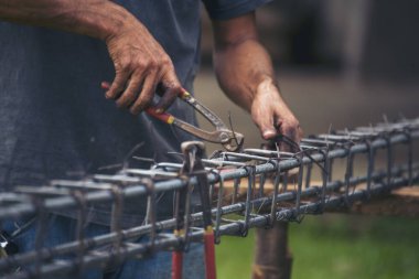 İnşaatçıların elleri çelik telleri büküyor beton işçiliğini destekliyor. Kerpeten kullanan işçi elleri demir tel kullanıyor. Açık hava işçisi, tel bükme pensesi kullanıyor, inşaat işi yapıyor.