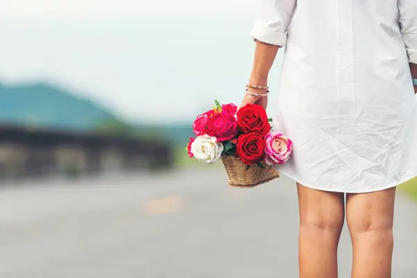 Woman hand holding rose flower hiding behind back side rear view to surprise couple on valentine\'s day. Beauty woman giving surprise red rose florist outdoor in green park. valentine\'s day concept