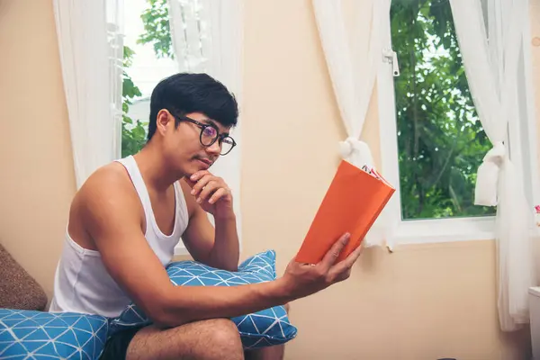 Relax man holding book read on sofa happiness. Young asian man reading book on cozy couch sofa at warm home. Man reading open book leisure mind in living room. Happy enjoyment man lifestyle at home