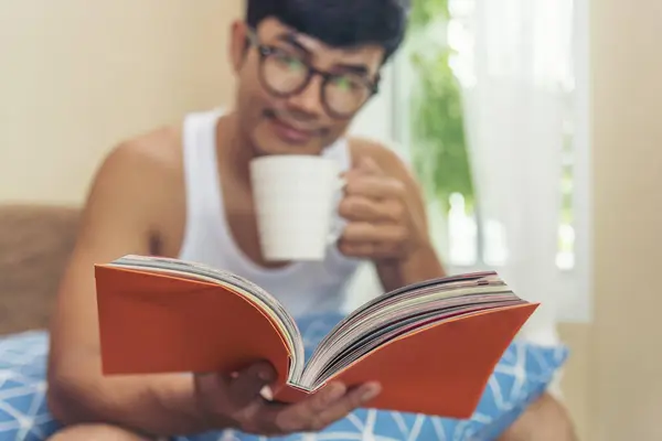 Young asian man reading book on cozy couch sofa at warm home. Relax man holding book read on sofa happiness. Man reading open book leisure mind in living room. Happy enjoyment man lifestyle at home