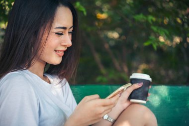 Asyalı kadın yeşil parkta tek kullanımlık sıcak kahve bardağı tutarken akıllı telefon kullanıyor. Banner Happy rahat Asyalı kadın açık hava bahçesinde gülümsüyor. Genç kadınlar fotokopi odasıyla doğanın tadını çıkarıyor