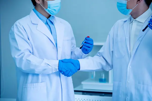 Two asian scientist men team partner shake hands together. Men researcher holding chemistry science tube experiment biotech antibody sample in laboratory. Close up hand corporate coworker partnership