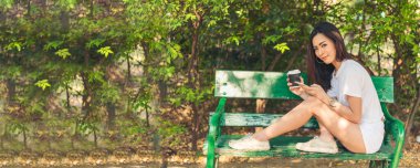 Banner Woman yeşil parkta elinde sıcak tek kullanımlık bardakla akıllı telefon içiyor. Web Afişi Mutlu Asyalı kadın açık hava bahçesinde gülümsüyor. Genç kadınlar fotokopi odasıyla doğanın tadını çıkarıyor