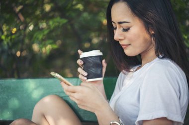 Asyalı kadın yeşil parkta tek kullanımlık sıcak kahve bardağı tutarken akıllı telefon kullanıyor. Banner Happy rahat Asyalı kadın açık hava bahçesinde gülümsüyor. Genç kadınlar fotokopi odasıyla doğanın tadını çıkarıyor