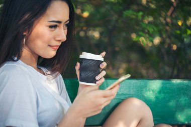 Asyalı kadın yeşil parkta tek kullanımlık sıcak kahve bardağı tutarken akıllı telefon kullanıyor. Banner Happy rahat Asyalı kadın açık hava bahçesinde gülümsüyor. Genç kadınlar fotokopi odasıyla doğanın tadını çıkarıyor