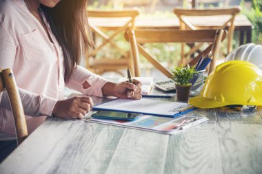 İnşaat mühendisi kadın, ofis masasında dizüstü bilgisayarla çalışıyor. Beyaz, inşaat ofisinde sarı kasketli. Asyalı genç adam mimarlık projesi yöneticisi ofiste oturuyor.