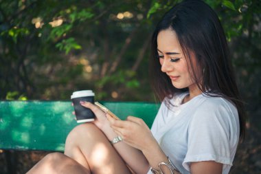 Asyalı kadın yeşil parkta tek kullanımlık sıcak kahve bardağı tutarken akıllı telefon kullanıyor. Banner Happy rahat Asyalı kadın açık hava bahçesinde gülümsüyor. Genç kadınlar fotokopi odasıyla doğanın tadını çıkarıyor