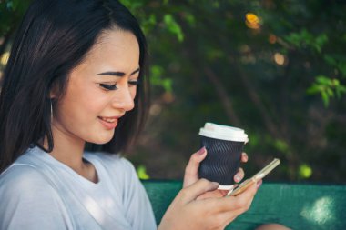 Asyalı kadın yeşil parkta tek kullanımlık sıcak kahve bardağı tutarken akıllı telefon kullanıyor. Banner Happy rahat Asyalı kadın açık hava bahçesinde gülümsüyor. Genç kadınlar fotokopi odasıyla doğanın tadını çıkarıyor