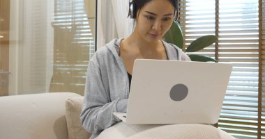 Mutlu Asyalı kadın video konferansı evdeki oturma odasında rahat koltukta oturuyor. Gülümseyen Asyalı kadın dizüstü bilgisayar kulaklığı kullanıyor. Evlerinden video izliyorlar.