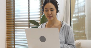 Mutlu Asyalı kadın video konferansı evdeki oturma odasında rahat koltukta oturuyor. Gülümseyen Asyalı kadın dizüstü bilgisayar kulaklığı kullanıyor. Evlerinden video izliyorlar.