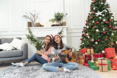 Mutlu Asyalı kadın Noel hediyesi kutusunda Noel ağacının altında birlikte akustik gitar çalıyor. İki kadın gülümsüyor. Oturma odasında Noel balosu, Noel ağacı, Noel bayramı, kış festivali konuşmaları.