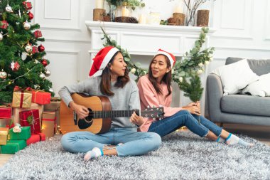 Mutlu Asyalı kadın Noel hediyesi kutusunda Noel ağacının altında birlikte akustik gitar çalıyor. İki kadın gülümsüyor. Oturma odasında Noel balosu, Noel ağacı, Noel bayramı, kış festivali konuşmaları.