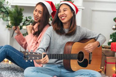 Mutlu Asyalı kadın Noel hediyesi kutusunda Noel ağacının altında birlikte akustik gitar çalıyor. İki kadın gülümsüyor. Oturma odasında Noel balosu, Noel ağacı, Noel bayramı, kış festivali konuşmaları.