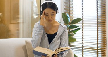 Stresli Asyalı kadın kulaklık takıyor. Oturma odasındaki rahat koltukta oturmuş müzik okuyor. Depresyondaki Asyalı kadın araştırma kitabında dergi okuyucusu baş ağrısı insanı okudu.